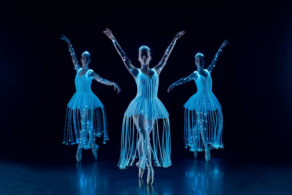 Tres bailarinas con trajes de ballet de luces LED sobre un escenario en penumbra